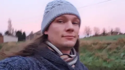 Family handout David Clark in a knitted hat and jumper. Behind him is a green field.