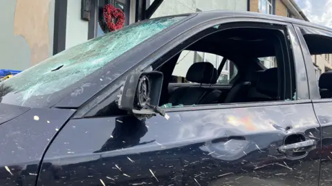 Shows a black car with its windows smashed and dents to the bodywork.