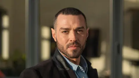 Willis has a shaved head. Wearing a black coat and denim shirt. He stands outside a floor to celling windows