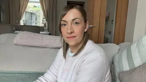 Sophie Dodd Trainee midwife Sophie Dodd. She is sat down on a sofa in a living room. She has mid length light brown hair and is wearing a pink cardigan.