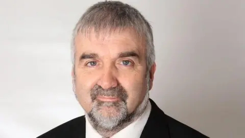 A man looking at the camera, he has a grey/black beard and grey hair