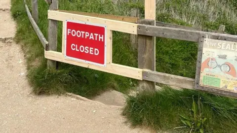 East Riding of Yorkshire Council A red sign with white writing that reads "Footpath closed" in capital letters is attached to a wooden fence that is blocking access to steps.