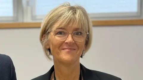 A woman with a blonde bob wearing a navy blazer and clear glasses smiling for the camera standing in an office room. The shoulder of another person is visible on the left.