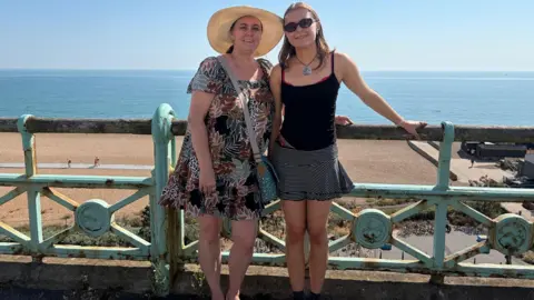 Caroline Herman Caroline (L) stood next to her daughter (R). They are leaning on an ornate railing with their backs to a beach looking out to the sea. Caroline is wearing a dress and large summer hat and her daughter is wearing a black top and a skirt.