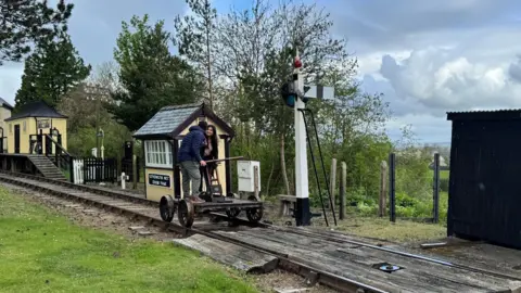 A man and a woman stand on a vintage push trolley which they are manually pushing along a railway track.