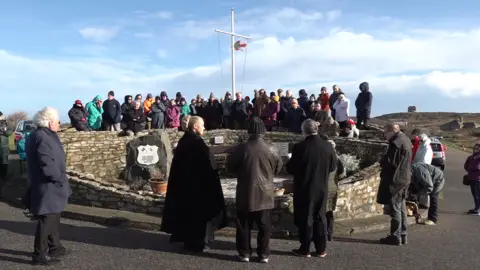 Crowds and spokespeople gather around the Hammond Memorial in Alderney. More than 30 people are stood around the stone memorial site. The sky is overcast. The Alderney flag is flying. 