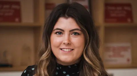 Electrical Safety First Karter Kane, a young woman with long, wavy hair, smiles at the camera.  She has blue eyes and her hair is dark on top with blonde highlights.  She is wearing a black belted dress patterned with light blue spots.  
