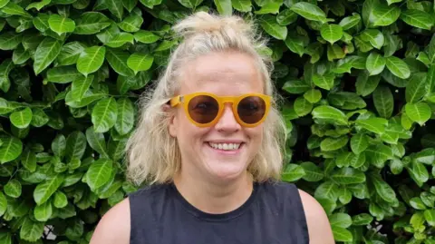 Jen Mansour is smiling and standing in front of a large green bush.
She is wearing yellow-rimmed sunglasses and a black top.
She has long blonde hair which is tied back.