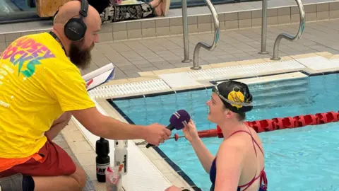 BBC Charlotte is standing in a swimming pool wearing a black Children in Need swimming cap. She is speaking into a purple microphone. A man with a bald head and long beard is wearing a yellow Children in Need T-shirt and is crouching on the side of the pool, holding the microphone to Charlotte's mouth.