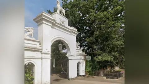Bourne Hall Museum in Ewell Village. The entrance has one large arch and two smaller arches either side. There is a dog at the top of the highest arch. A big tree can be seen to the right of the entrance, with a sign stating 'Bourne Hall'.