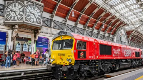 A red train with '200' written on the side at a platform at Paddington Station. There is a large station clock to the left and passengers waiting to get on the train in the background