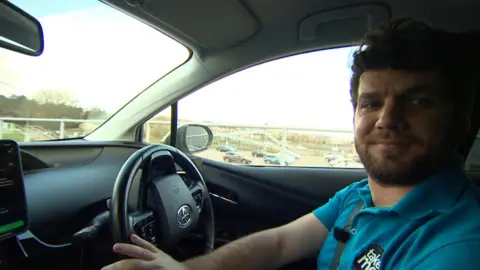 Julian Pitorac smiles off camera in a blue polo shirt with a 'TakeMe' badge. He is sat in the drivers seat of a car and is turning the steering wheel.