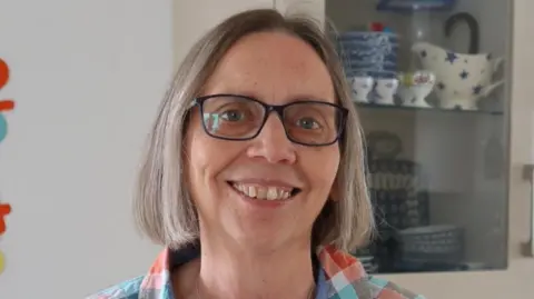 Hilary Stobart is smiling at the camera, standing in front of a display cabinet full of china. She has bob-length fair or greying hair and is wearing dark-rimmed spectacles and a colourful checked shirt