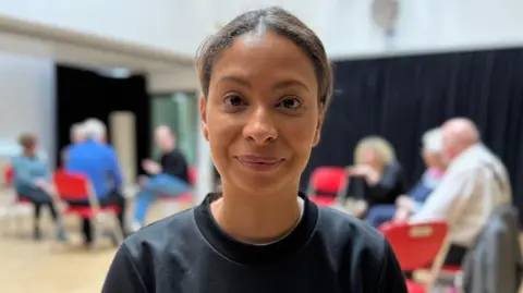 A young, black woman with pinned back hair wearing a black round neck jumper.