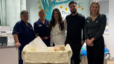Manx Care Parents Hannah Spearing and Matt Knight are standing with the staff at the maternity unit at Noble's hospital, and a woman from the Abigail's Footsteps charity. They're standing together in a ward behind a cot covered in blankets with a teddy bear inside. You can see a tree mural behind them on the ward's wall.
