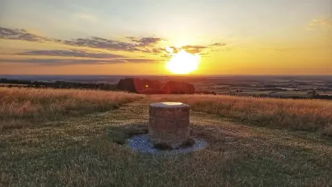 Dotty A stone cylinder sits in the middle of the foreground, possibly with a metal distance map on top, with the countryside, fields and lines of trees, spreading out behind it. A large yellow sunset fills the top third of the picture.