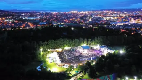 Féile an Phobail Crowds at a night concert at Falls Park. There are trees beside the venue and bright lights around it