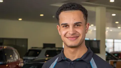 Tyrell Cambridge has short brown hair and is wearing a navy and silver polo shirt. He is standing in a car showroom which has several vehicles and large windows.