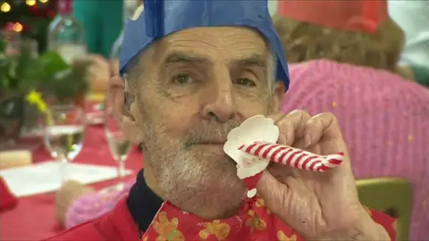 BBC A man is blowing a party blower that is red and white. He is wearing a blue Christmas cracker hat and is wearing a red jumper.