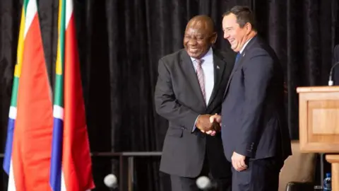 Gallo Images/Getty Images A smiling President Cyril Ramaphosa in a grey pinstripe suit shakes hands with John Steenhuisen of the Democratic Alliance - who is also smiling and in a black suit. South African flags can be seen to the left of the shot.