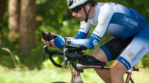 Mark Beaumont A man in a blue and white lycra cycling outfit rides a bike through a woodland.