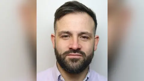 Gloucestershire Constabulary Police custody picture of Mark Bale. He is wearing a purple shirt and has a beard and dark hair combed to the side. 