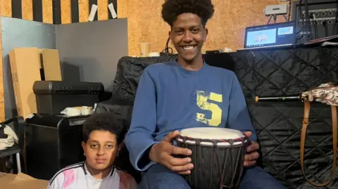 Two boys sitting next to each other. One is wearing a pink and white shirt and the other is wearing a blue shirt while holding a drum.