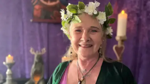 Priestess Sarah Robinson smiling with a green and white flower gardland in her hair and wearing nose rings and multiple necklaces with a purple fabric in the background and some candles. 