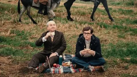 Luis Gnecco in brown suit and Fionn Whitehead in dark duffel coat are having a picnic, sitting on a rug in a field. The lower legs of two donkeys can be seen in the background.