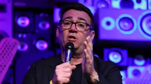 EPA/Shutterstock Manchester Mayor Andy Burnham speaking on a microphone with music speakers behind