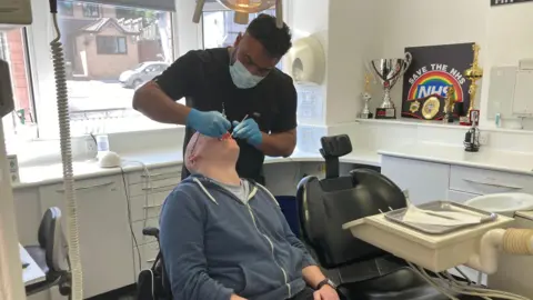 A man in a blue hoodie has his mouth open and is being examined by a dentist, wearing a mask and gloves and a black top.  On the wall of the examination room is a sign with 'save the NHS' and a number of trophies and boxing belt.