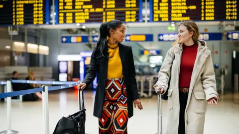 Two ladies carry their suitcases through an airport with a full departure board behind them