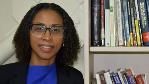 Anselm Gibbs Dr Malisa Neptune-Figaro, wearing glasses and a blue shirt and black jacket, stands next to a book shelf at the University of the West Indies 