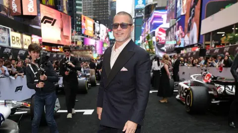 Getty Images Brad Pitt stands in a black suit in front of multiple F1 racing cars in New York City at a promotional event for his film F1. Electonic billboards can be seen behind him on the streets of Times Square - including one for F1. 