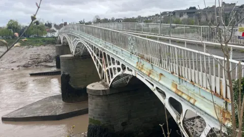 The image shows an empty bridge crossing a muddy brown river, with exposed banks on either side. The bridge is painted light blue, with visible rust along its sides. In the distance, a town can be seen with an old castle.