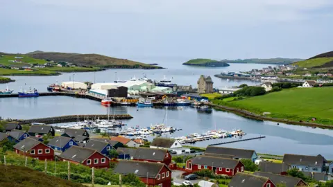 Getty Images An image of the port town of Scalloway in the Shetland Islands - green hills rise from the blue waters, with red houses scattered around the bay where lines of boats are moored