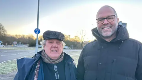 A man in a navy jacket, tartan scarf, and black flat cap stands roadside by a man in a black hooded puffer jacket with glasses. 