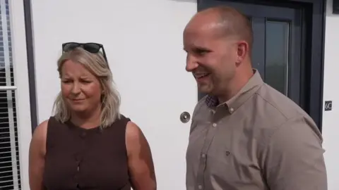 Jess and Nick looking to the left and smiling in front of their newly built home. He is wearing a brown shirt, and she's wearing a black top with sunglasses on her head.