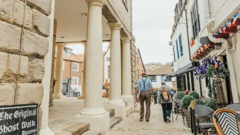 North Yorkshire Council A cobblestone street lined with historic buildings, including a stone structure with large columns on the left and white‑painted shops decorated with red, white, and blue bunting on the right. Patterned outdoor café chairs and potted purple flowers sit along the walkway as people stroll through the street.