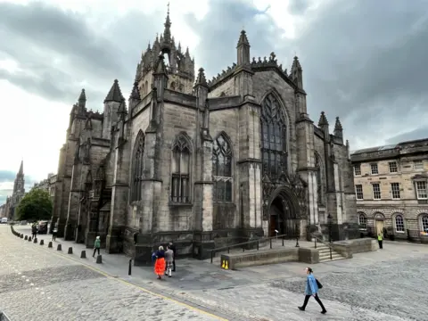 St Giles' Cathedral in Edinburgh, taken from across the Royal Mile, facing east. The west front of the cathedral can be seen under a grey sky. Five people are walking by in a scattered fashion.