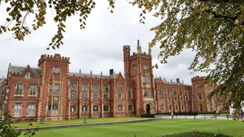 PA Media A photo of Queen's University main building (the Lanyon building), it's red bricked with smaller yellow masonry around the windows, which consist of 3 floors of windows and larger windows on the top floor. A central tower is the main entrance with four floors of windows. The building sits on a patch of green grass with a walkway to the main entrance. 