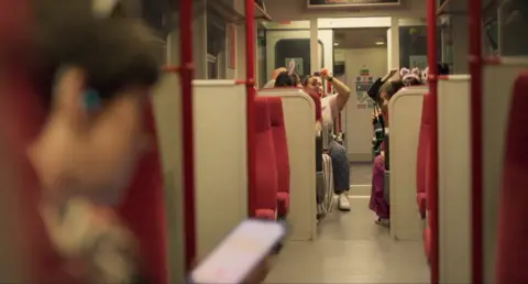 Euston Films/BBC A group of passengers can be seen at one end of a train carriage. In the foreground a woman looks at a mobile phone screen. She is out of focus.