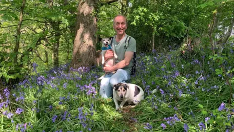 Pete is sitting in woodland filled with purple wildflowers. He is holding his dog in his arms with his second pooch sitting next to him. He is smiling directly at the camera.