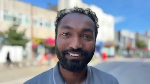 A man with a beard and short dark hair smiling at the camera. He's smiling and stood on a quiet high street wearing a blue polo top. 