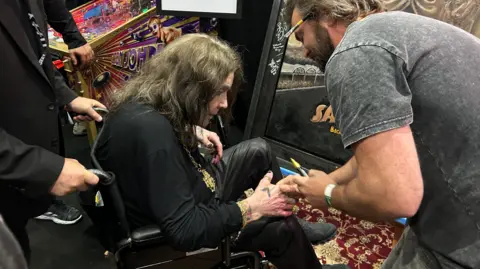 Ozzy Osbourne, in a wheelchair, is shaking hands with a man who is stood up. The man is holding a yellow marker pen.