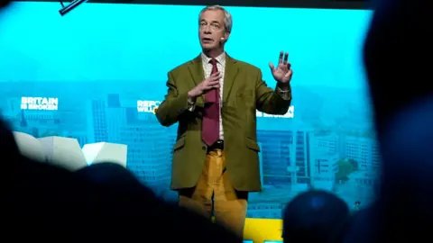 Peter Byrne/PA Wire Nigel Farage stands in front of a blue screen saying "Britain is broken". In the foreground can be seen silhouettes of the audience.