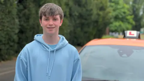 BBC Alex has straight, brown hair and wears a light blue hoodie. He stands next to a driving school car.