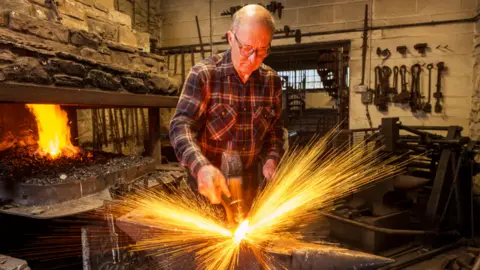 Mark Lewis Paul Dennis is an older man with a balding head and a grey fringe of hair. He wears glasses and a checked workman's shirt. He is in his workshop with a lit coal forge to the left of the picture. He is hammering something on an anvil. The photograph is done in long exposure so there are streaks of sparks making a sort of firework effect around the metal being worked. There are lots of metal tools and other machines in the background.