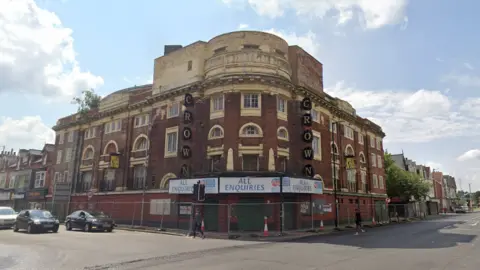 Google Google Maps image of the former Crown pub. It is a red-brick building with discoloured and peeling cream paintwork around the many windows. It stands on the corner of the street and is surrounded by metal fencing. Sale signs are above the main entrance. The pub's name is spelled out on either side of the corner section in huge black round signs vertically alligned and each containing a single letter. From top to bottom they spell out CROWN.