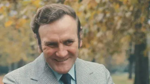 Don Revie with combed brown hair, smiling, as he walks in a park. He is wearing a grey blazer, blue shirt and navy tie. There are autumn trees behind him. 